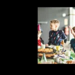 Children wearing party hats are sitting at a table decorated with a green tablecloth, filled with various food items including pizzas and desserts. The excitement is palpable at the football birthday party, with one child enthusiastically raising their hand. The background shows a blurred indoor setting. © Powerleague