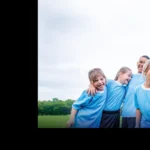 A group of young children in light blue jerseys stand closely together on a grassy field, laughing and smiling. The sky is overcast, and trees are visible in the background. This image from one of the football holiday camps is framed to the right, leaving a black area on the left side. © Powerleague