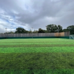 A football field with green artificial turf, perfect for local football leagues, features white goalposts. It's encircled by a high wooden fence and tall trees. The sky is overcast with patches of blue peeking through the clouds. © Powerleague