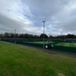 An outdoor sports complex with several fenced artificial turf soccer fields hosts local football leagues under a cloudy sky. Numbered signs are visible on the fences, and the foreground shows grassy areas with picnic tables to the left. © Powerleague