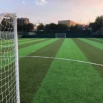 A soccer field with green and darker green striped artificial turf hosts local football leagues. A goalpost and net are in the foreground, with another in the distance. The sky is clear, and trees and buildings dot the background, setting a perfect scene for grassroots competition. © Powerleague