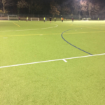 A night view of a soccer match in progress on a well-lit artificial turf field. Players in neon and dark uniforms, representing local football leagues, are scattered across the field, with a goalpost visible in the background. Trees surround the field, and shadows are cast by the bright stadium lights. © Powerleague