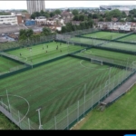 Aerial view reveals multiple fenced soccer fields with vibrant green artificial turf, hosting players likely from local football leagues. Surrounding the pitches are residential houses, industrial buildings, and a large sports stadium looming in the background. © Powerleague