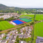 Aerial view of a sports complex, where local football leagues play on vibrant blue and green fields for hockey, enclosed by red and blue fences. The surrounding residential area features houses, roads, lush greenery, and hills in the background under a clear, bright sky. © Powerleague