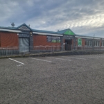 A one-story brick building with a green roof and signs, including a gym logo, stands empty beneath the cloudy sky. The parking lot is deserted, save for a person near the entrance, perhaps contemplating joining as part of their local football leagues' training regimen. © Powerleague