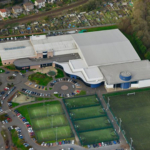 An aerial view of a sports complex featuring multiple buildings with large roofs, surrounded by parking areas with numerous cars. The complex includes several green tennis courts and a large synthetic turf field, frequently used by local football leagues. Residential houses and gardens are visible nearby. © Powerleague