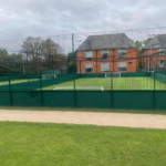 A fenced outdoor sports field with artificial grass, featuring two small soccer goals, serves as a lively venue for local football leagues. The background showcases a red brick building with several windows and a gray roof. The sky is overcast, with a grassy area in the foreground adding charm. © Powerleague