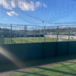 A sunny outdoor tennis court, enclosed by a green fence and netting, echoes the community spirit found in local football leagues. The court boasts a logo on its side barriers and is framed by trees under a clear blue sky, with sunlight streaming in from the left. © Powerleague