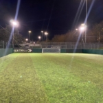 A well-lit outdoor soccer field at night, featuring a green turf pitch surrounded by high netting and fencing. The goalpost is positioned at the far end, and lights on poles illuminate the playing area, perfect for local football leagues. Trees and greenery are visible in the background. © Powerleague