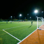 A nighttime soccer match unfolds on a well-lit artificial turf field, part of the vibrant local football leagues. Players are visible in the distance, actively engaged in the game. The sky is dark, and tall floodlights illuminate the scene, casting bright light onto the field. © Powerleague