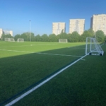 A soccer field under a clear blue sky hosts thrilling local football leagues, with several white goalposts placed on the green artificial turf. Tall residential buildings and a line of trees are visible in the background, as shadows stretch across the field. © Powerleague