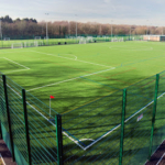 A large, artificial turf sports field hosts local football leagues, surrounded by a tall green fence. The field is marked with white and red lines for various sports and has goals on either end. Trees and a clear blue sky paint the backdrop. © Powerleague