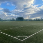 A photograph of an empty soccer field under a partly cloudy blue sky. The artificial turf field, used by local football leagues, has white boundary lines and several goalposts in the distance. Trees and tall streetlights surround the field, with a city skyline visible on the horizon. © Powerleague