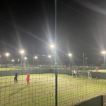 A nighttime soccer match from local football leagues is taking place on multiple illuminated fields, with players in jerseys actively playing. The fields are enclosed by netting, and bright floodlights cast a glow over the scene. © Powerleague