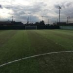 A small, enclosed soccer field with artificial turf under a cloudy sky serves as the perfect venue for local football leagues. The field is marked for gameplay, and a single goal stands at one end, illuminated by floodlights positioned around the perimeter. © Powerleague