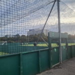 A fenced football field with artificial turf is shown, where local football leagues often play. Two people are walking near the gate, while trees and a building stand in the background under a cloudy sky. © Powerleague