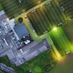 Aerial view of a sports complex with illuminated soccer fields hosting local football leagues at night. The facility includes a parking area with cars, a main building, and several well-lit, rectangular fields surrounded by greenery. © Powerleague