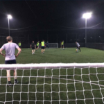 People enjoying a night-time soccer match on a small, well-lit field, part of their local football league. One player stands near a goalpost while others, in neon bibs, move across the field. The scene is enclosed by a netted fence. © Powerleague