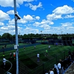 A group of people play soccer on a green outdoor field under a bright blue sky with scattered clouds, showcasing the spirit of local football leagues. A crowd watches from the sideline behind a fence, while several smaller fields are visible in the background. © Powerleague