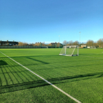 A spacious soccer field with artificial green turf, two small goals, and a clear blue sky hosts local football leagues. The field is surrounded by a fence, and residential buildings are visible in the background. © Powerleague