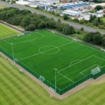 Aerial view of a sports complex featuring two adjacent soccer fields, home to local football leagues, surrounded by greenery and trees. One field is enclosed with fencing and has goalposts. Nearby, a road winds past industrial buildings in the background. © Powerleague